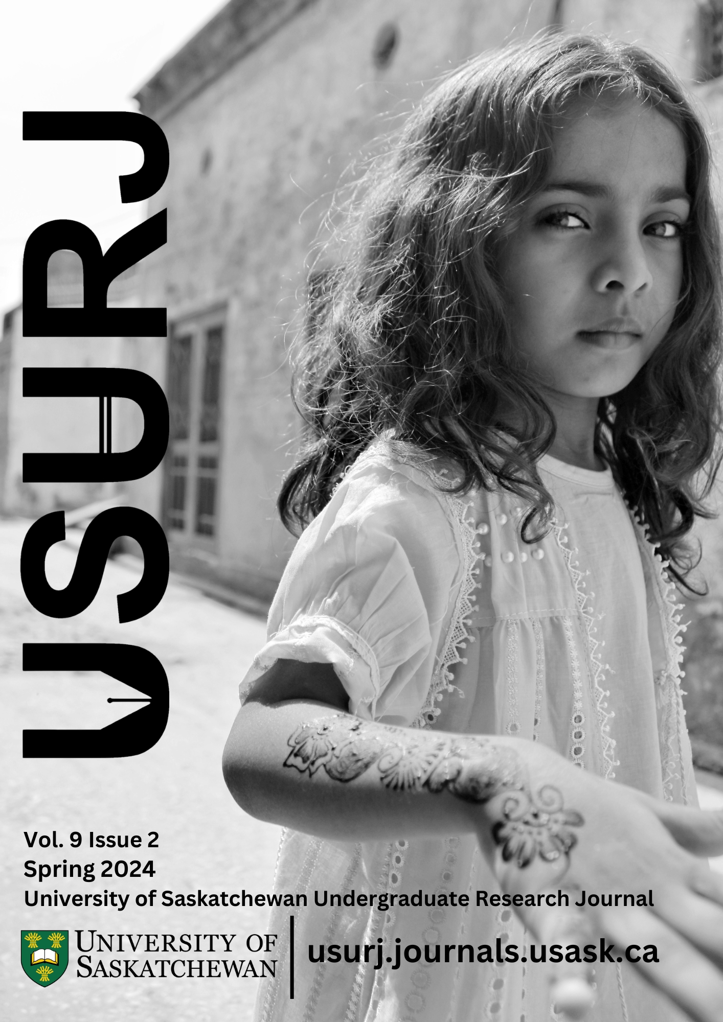black and white photograph of a young girl in Pakistan, with long wavy hair, a confident stare, and white cotton dress. Her forearm and hand have intricate henna patterns. She takes up most of the frame, standing in a street near a building.